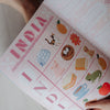 A child's educational poster or activity book page titled 'INDIA' being held open by hands, showing colorful illustrations of Indian symbols, animals, and objects on a light pink background.