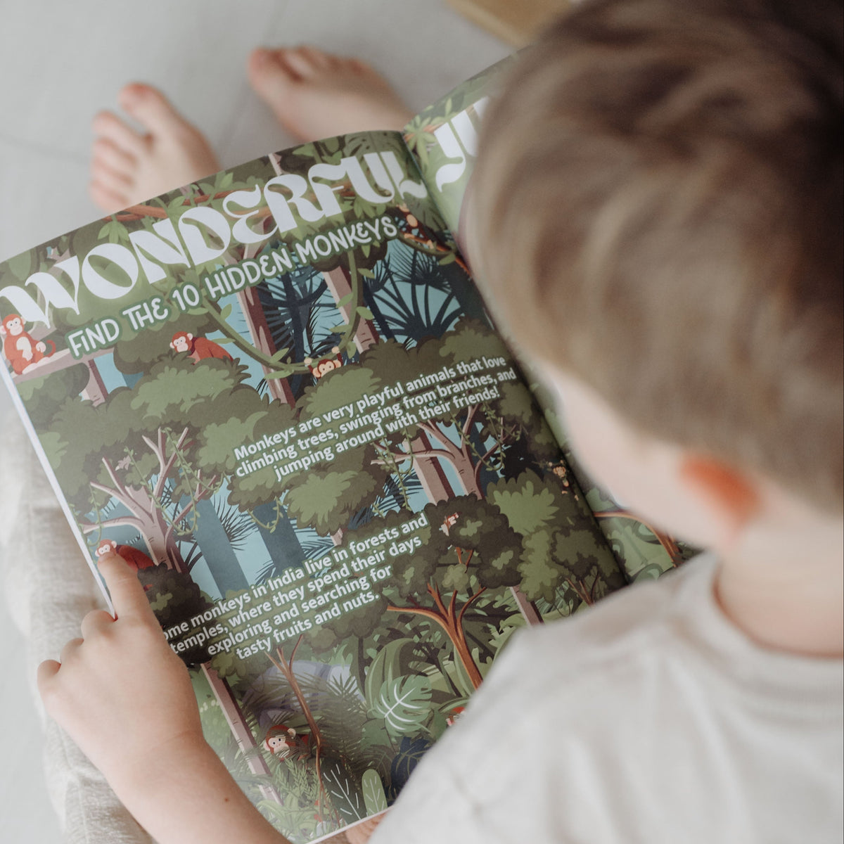 A child sitting on a beige cushioned floor mat, holding a book titled 'Wonderlust' in a bright, minimalist room with white wooden floors.
