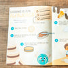 An open children's cooking book spread on a wooden surface, featuring illustrated steps for making alfajores with colorful drawings, photos of cookies, and cartoon characters.