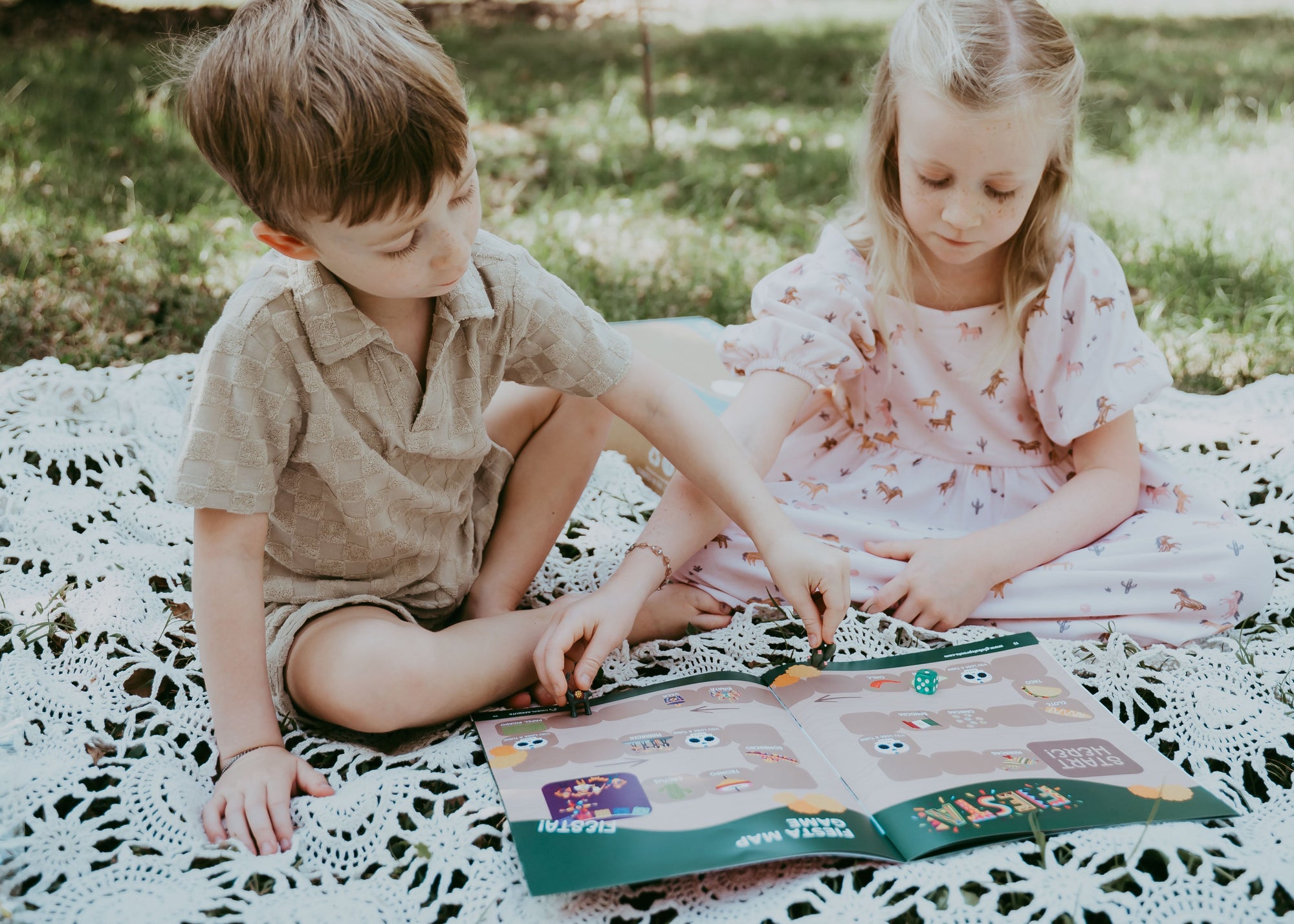 Two children sitting on a blanket outdoors, reading a book.